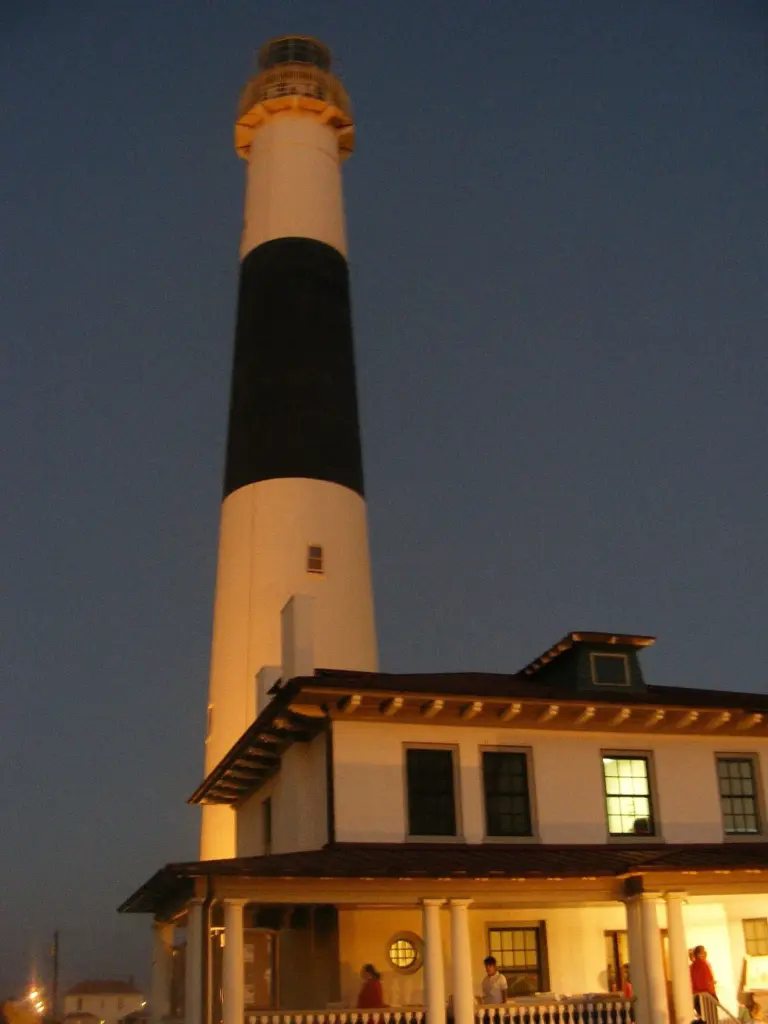 A tall lighthouse tower with a black middle band, lit at dusk above a keeper's house with glowing windows