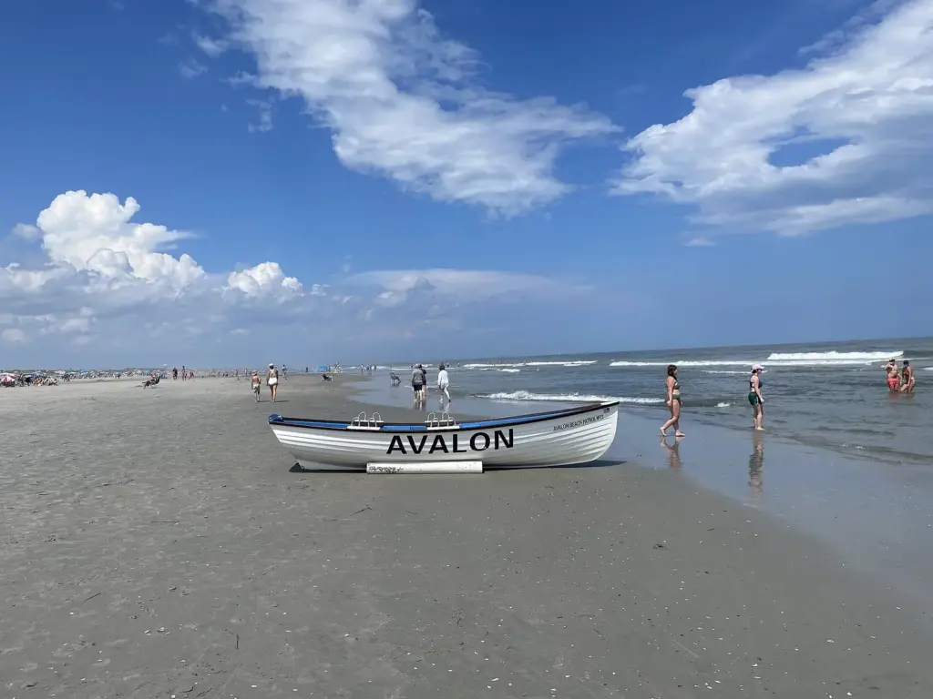 A white Avalon Beach Patrol lifeboat on the sand at Avalon, with swimmers in the surf under a blue sky