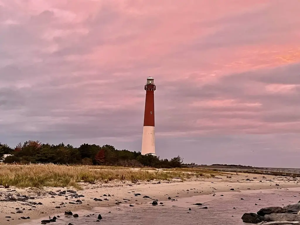 Barnegat Lighthouse, a red and white brick tower, rising behind the beach at Barnegat Light under a pink dawn sky