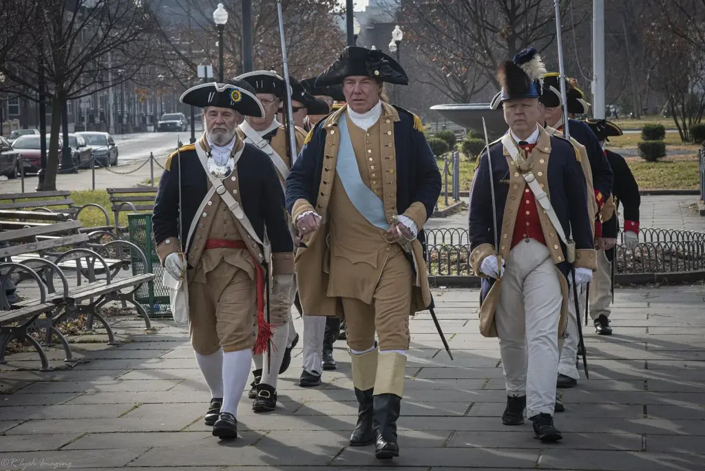 Reenactors in Continental Army uniforms and tricorn hats marching along a park path lined with benches on a winter day