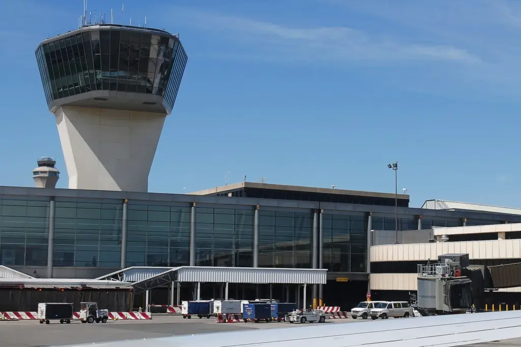 A concrete air traffic control tower with a wide glass cab rising above a low terminal building and parked service vehicles