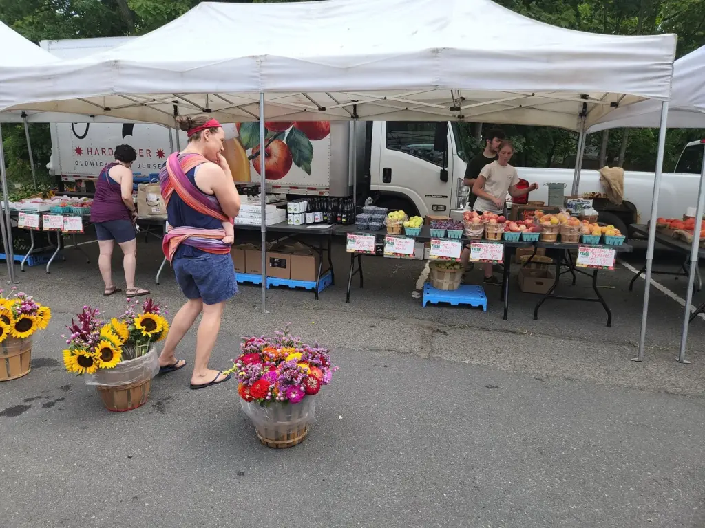 Shoppers browsing produce, peaches and buckets of sunflowers under white tents at a New Jersey farmers market
