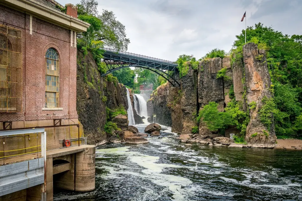 The Great Falls of the Passaic River pouring into a rocky gorge beside a brick hydroelectric plant in Paterson, with a steel arch bridge above