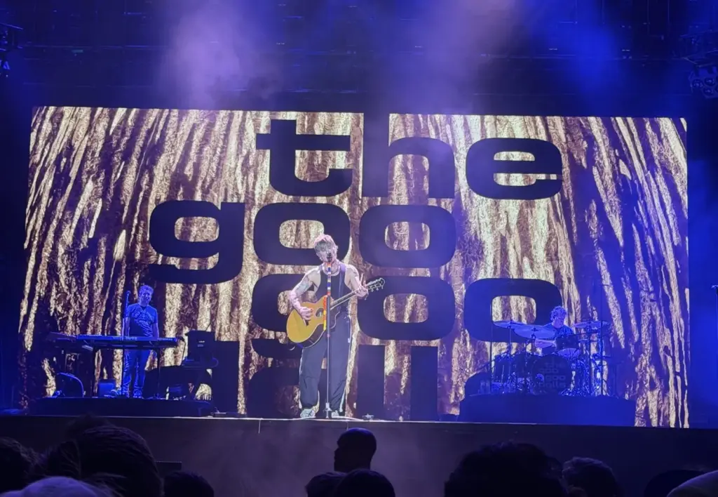 A band performing on a festival stage in front of a giant video screen, with the crowd's heads silhouetted in the foreground