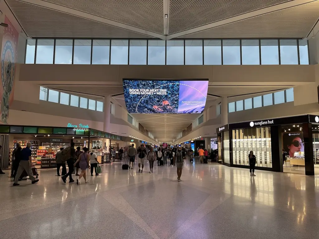 A long, high-ceilinged terminal concourse lined with shops and a large overhead video screen, with travelers pulling suitcases