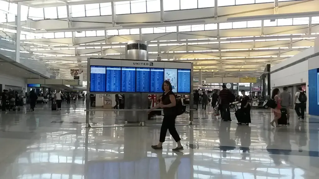 A bright, glass-walled airport departure hall with rows of blue flight information screens and travelers walking across a polished floor