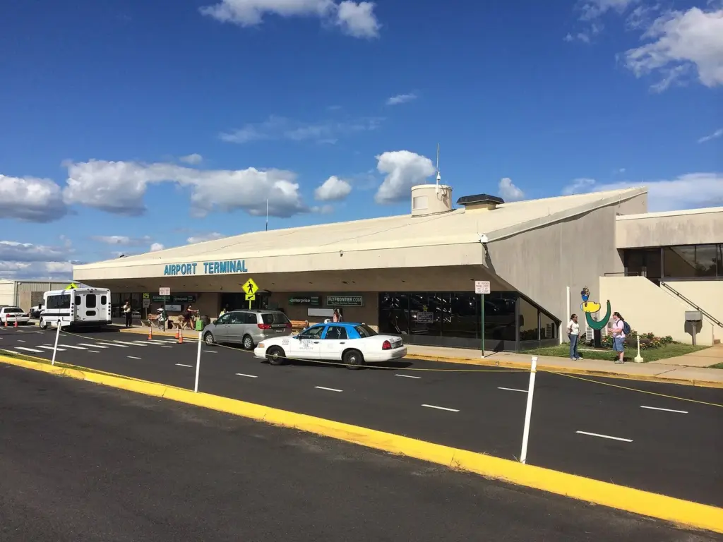 A low, 1970s-style airport terminal building with a sign reading Airport Terminal, a taxi at the curb and a blue sky