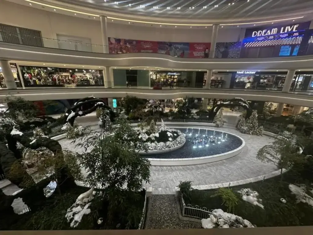 A landscaped indoor court with trees, rocks and a round fountain, ringed by shopping balconies, with a lit Dream Live sign on the upper level