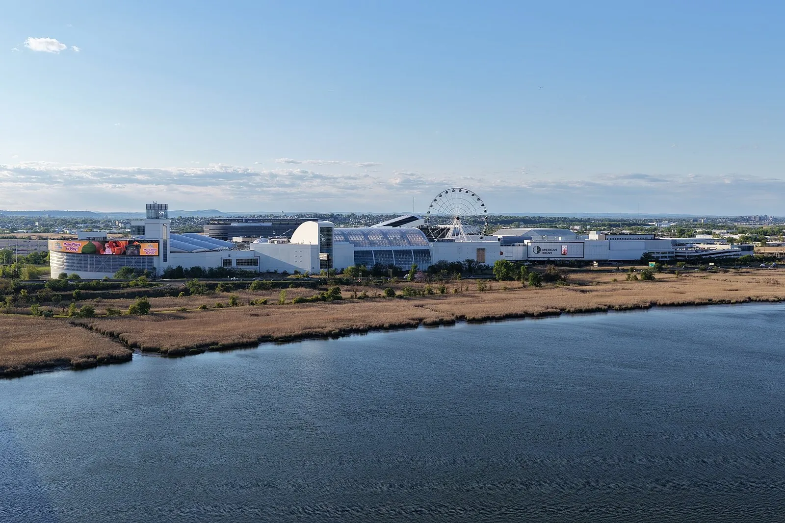 The American Dream mall from the air, a long white complex with glass-vaulted halls and the Dream Wheel, with MetLife Stadium behind it and Meadowlands marsh and water in front