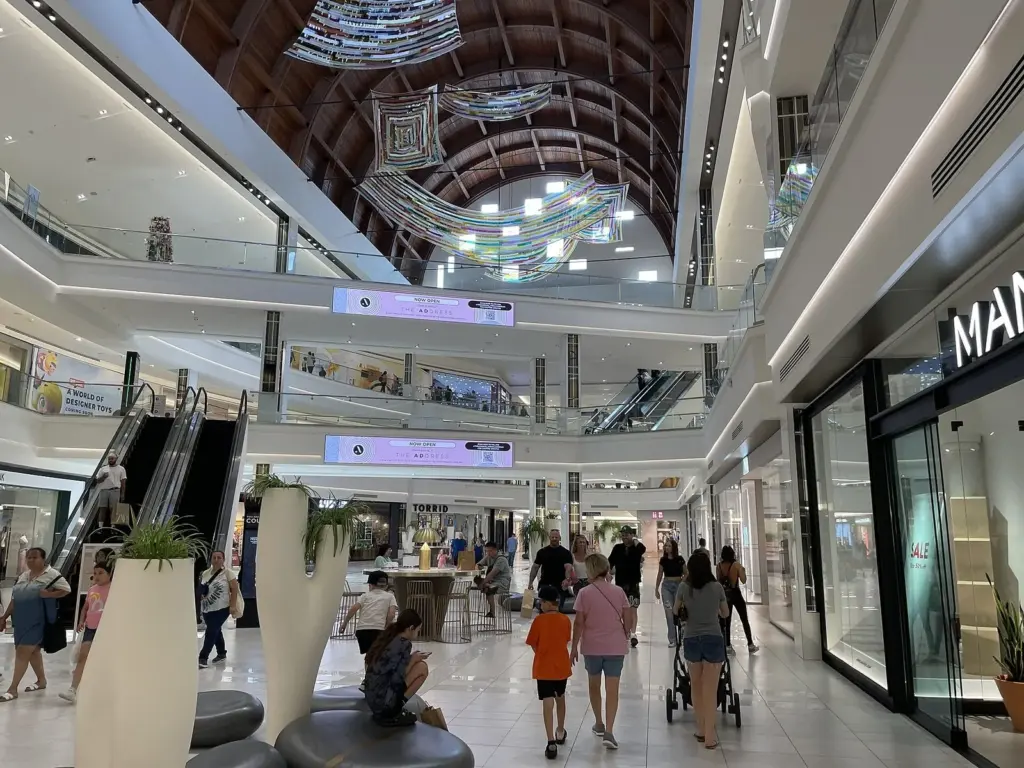 Shoppers walking along a bright first-floor concourse lined with stores, escalators and glass railings under a curved wooden ceiling