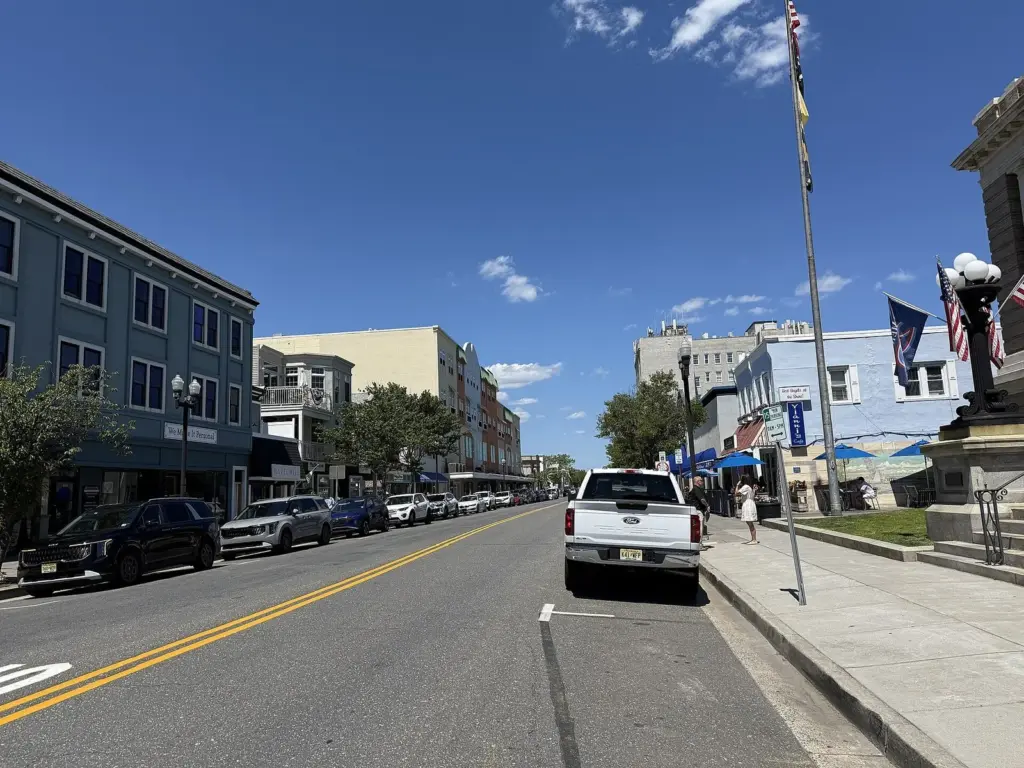 Asbury Avenue on a sunny day, a two-lane downtown street lined with two- and three-story storefronts and parked cars, with blue cafe umbrellas and flags on the right