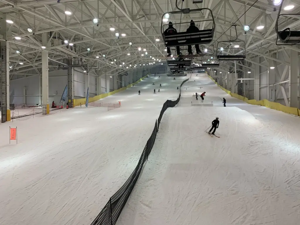 Skiers and snowboarders on a long white indoor slope under bright lights, with chairlift seats overhead and a dark safety net running down the middle of the run