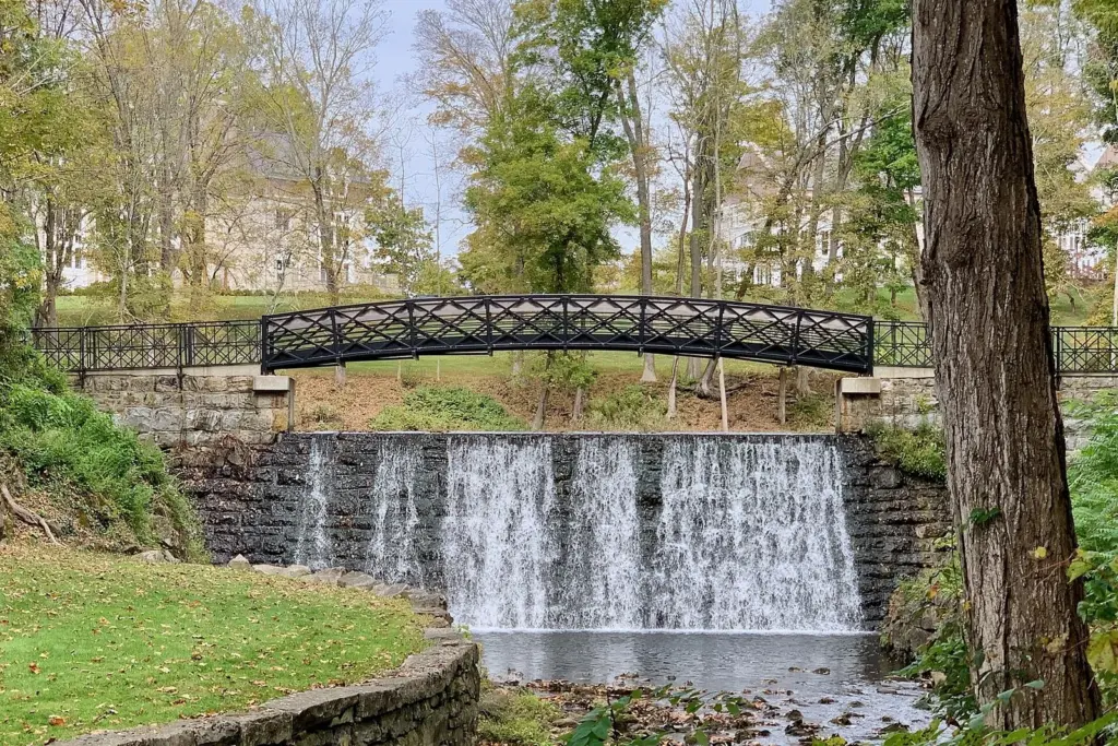A stone dam with water spilling over it beneath a black iron footbridge, framed by trees turning yellow in early fall