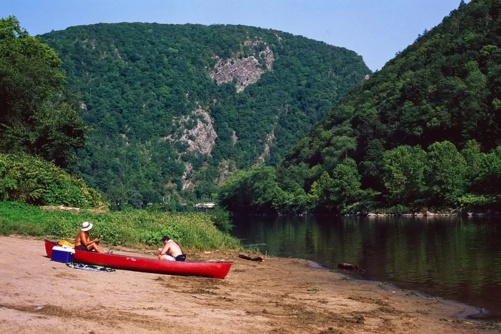 Two people resting beside a red canoe on a sandy riverbank, with the steep forested walls of the Gap rising behind the calm river