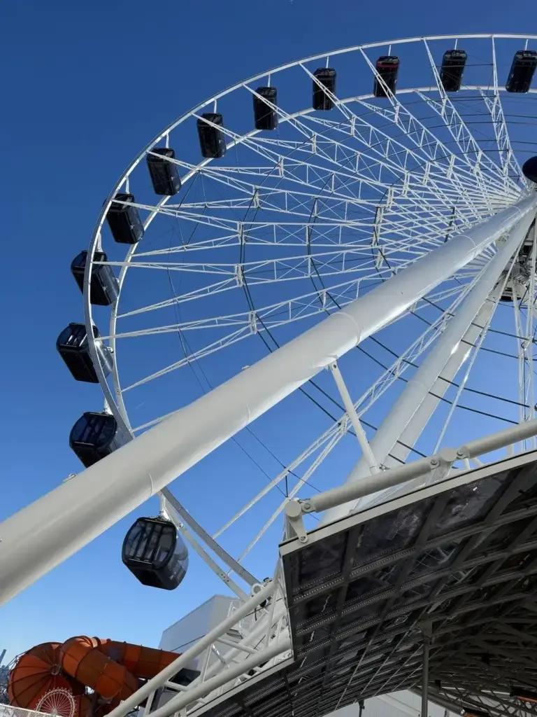 A white observation wheel with dark enclosed gondolas, seen from directly below against a clear blue sky