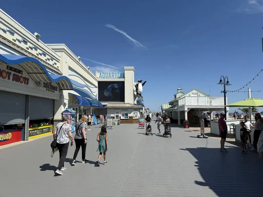 Families walking along a wide boardwalk lined with arcades and a building with an aquarium sign under a bright blue sky