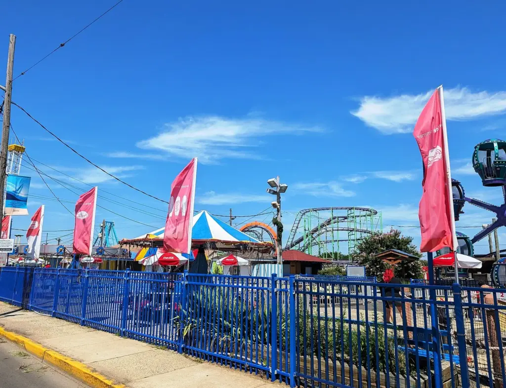 Colorful flags, a striped carnival tent and a roller coaster rising behind a blue fence at Keansburg Amusement Park on a sunny day