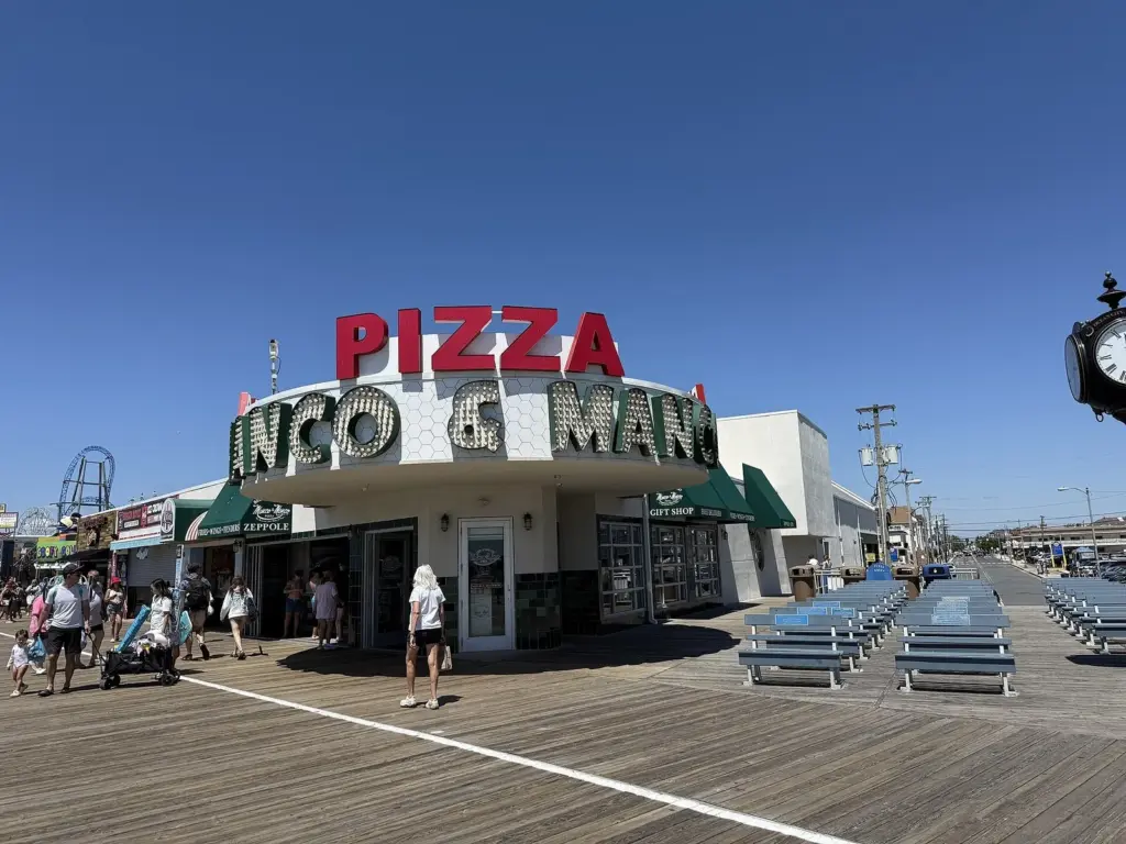Manco and Manco Pizza's round corner storefront on the boardwalk, with a tall red PIZZA sign on the roof and rows of benches out front
