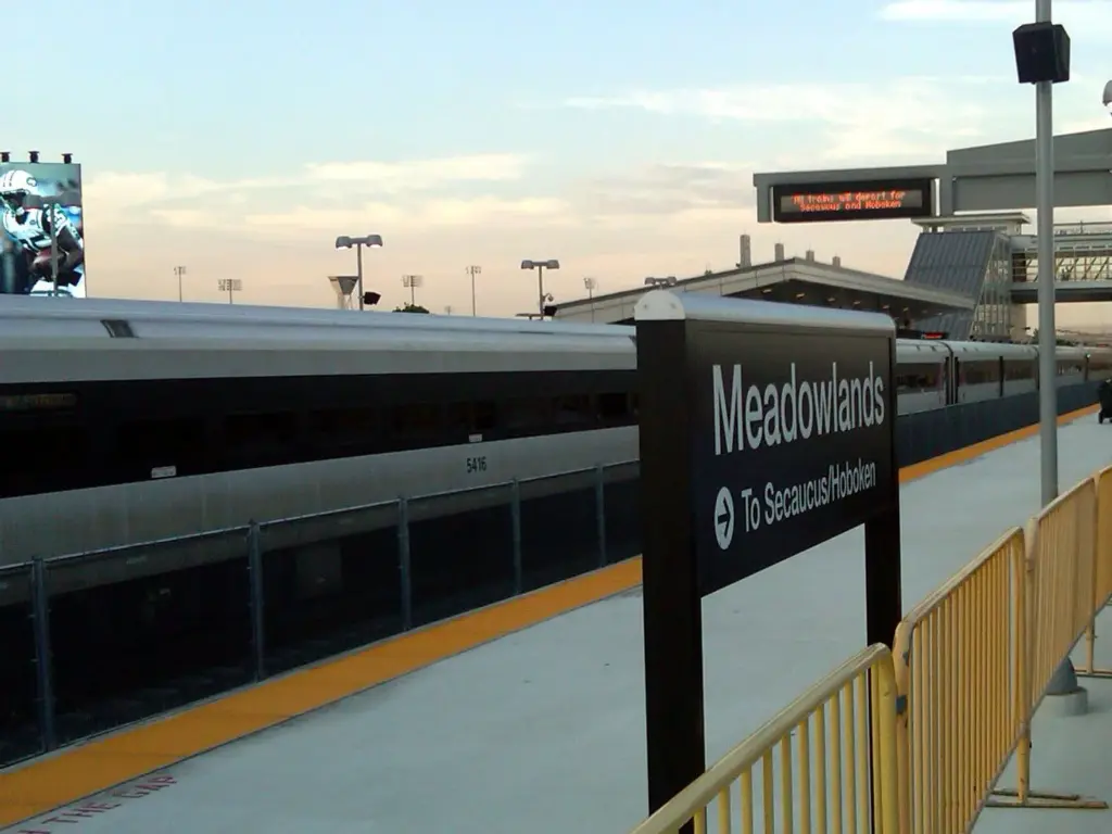 A double-decker NJ Transit train at the Meadowlands station platform, with a black station sign pointing to Secaucus and Hoboken and crowd barriers along the edge