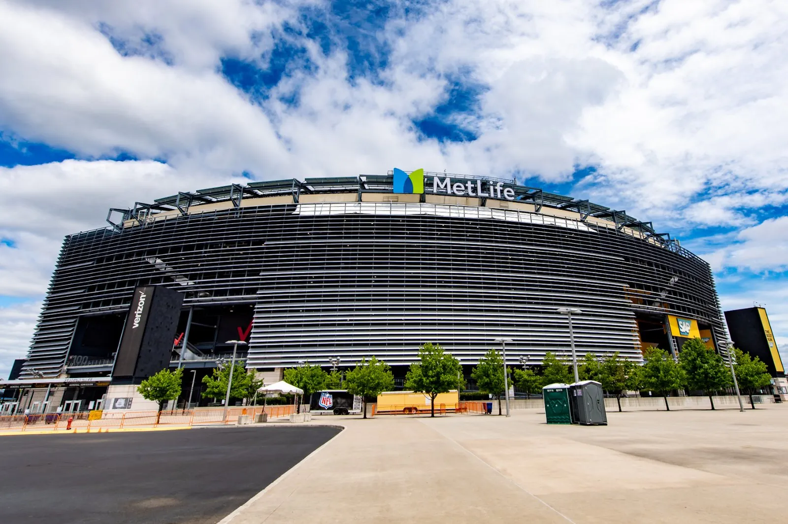 The gray louvered outer wall of MetLife Stadium with its blue and green MetLife sign, seen across the stadium plaza under a blue sky with scattered clouds