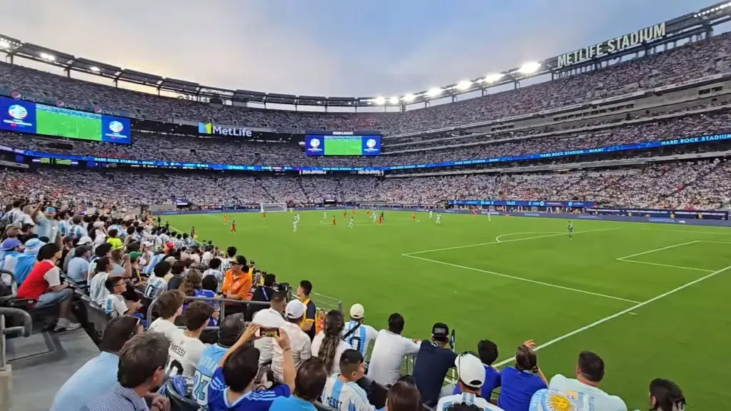 Fans in Argentina shirts in the lower bowl watching a soccer match, with the stadium's three tiers of seats and video boards across the field