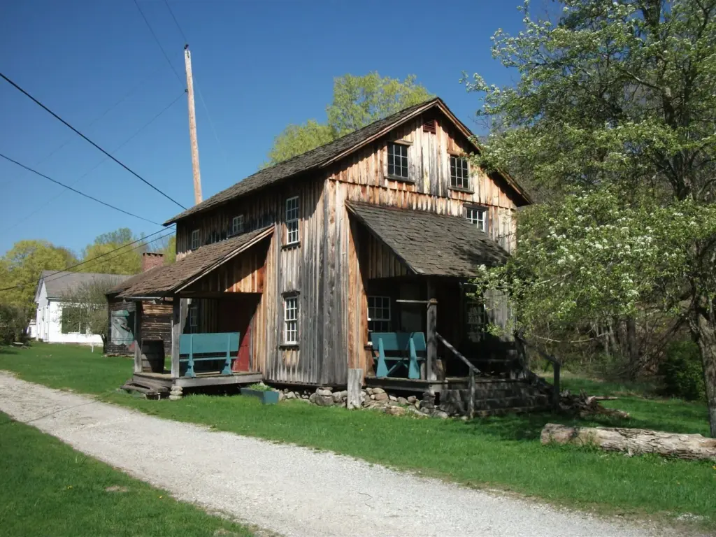 A weathered two-story wooden building with a shingled roof and a small porch beside a gravel lane at Millbrook Village, surrounded by spring trees