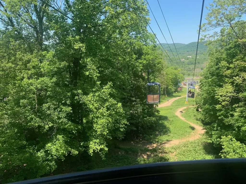 View from an open lift chair climbing a green ski slope in summer, with a dirt bike trail winding below and the Vernon Valley hills beyond
