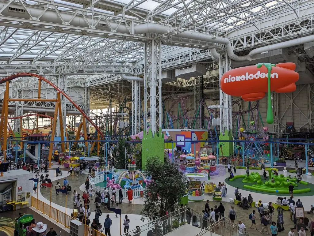 Nickelodeon Universe seen from an upper level, with orange roller coaster track, ride platforms, crowds of visitors and an orange Nickelodeon blimp hanging under the glass roof