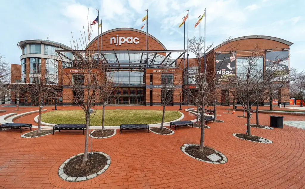 The curved red-brick front of the New Jersey Performing Arts Center, with flags on the roofline and a paved plaza ringed by young trees and benches