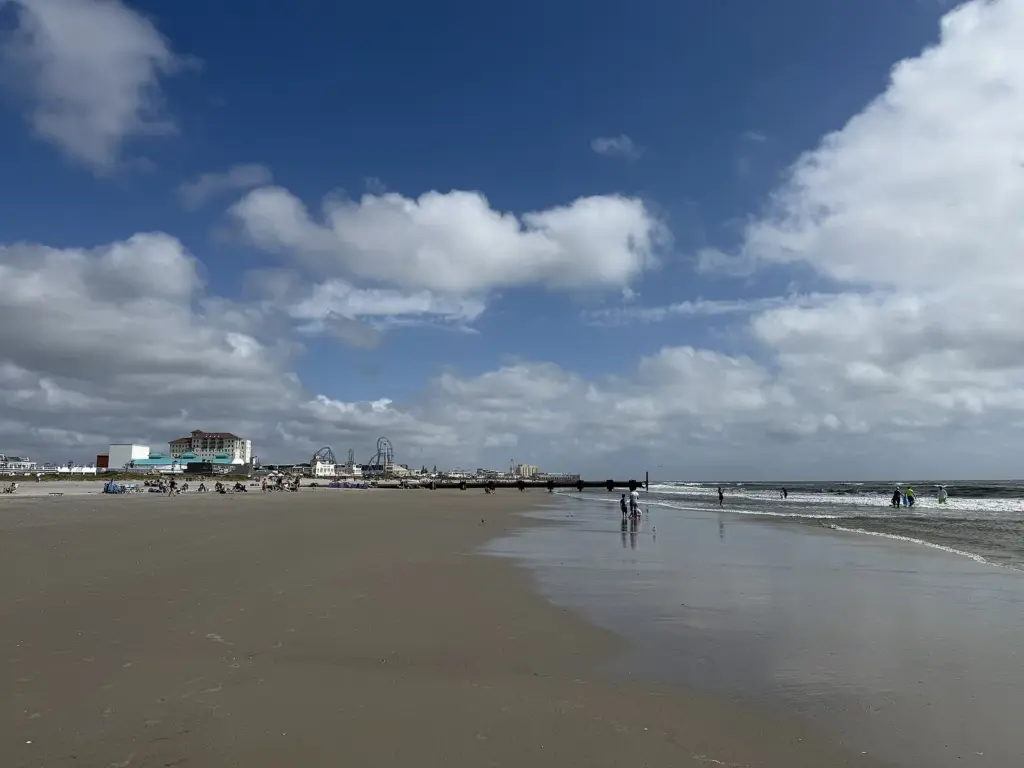 A broad, nearly empty stretch of Ocean City beach at 14th Street under big clouds, with a few people in the surf and hotels and roller-coaster tracks on the skyline