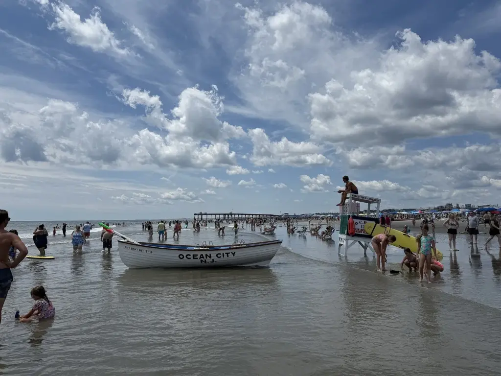 A lifeguard on a tall white stand marked Watch Your Children, with a white wooden lifeboat lettered with the town's name in the shallows and families wading around it