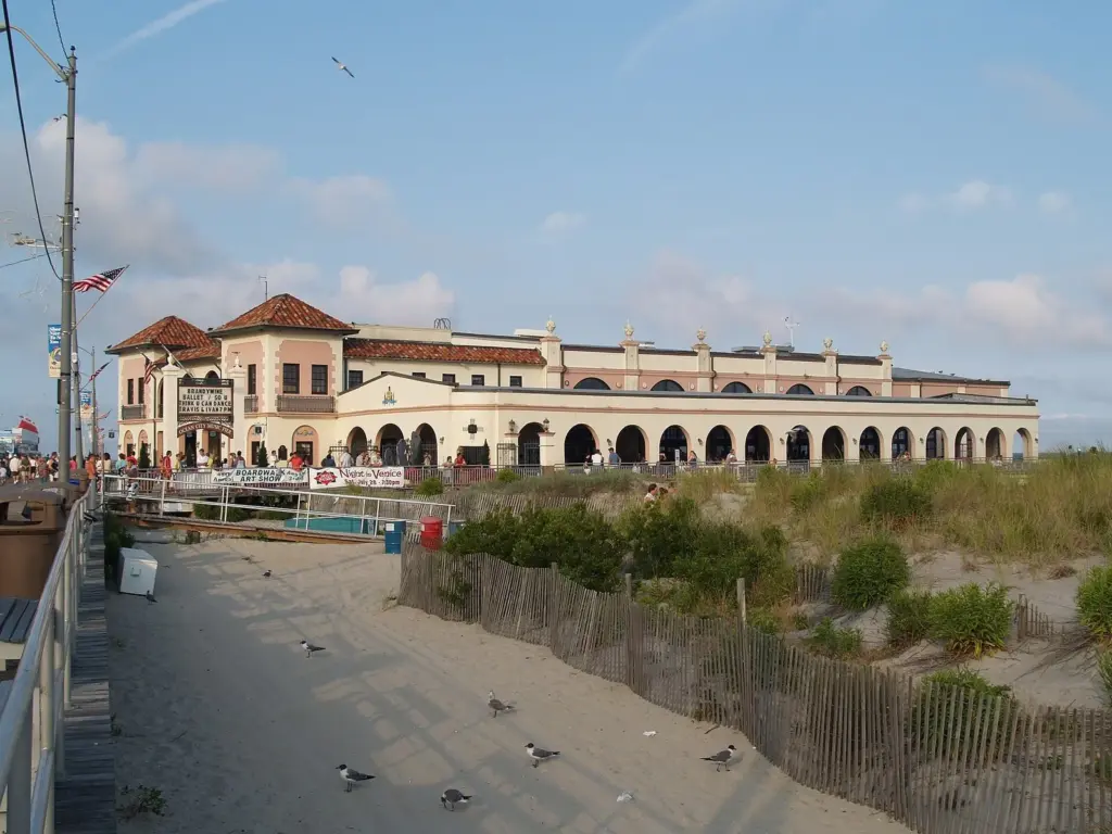 The Music Pier, a long cream-colored Spanish-style building with red tile roofs and a row of arches, built on the beach beside the boardwalk with dunes and fencing in front