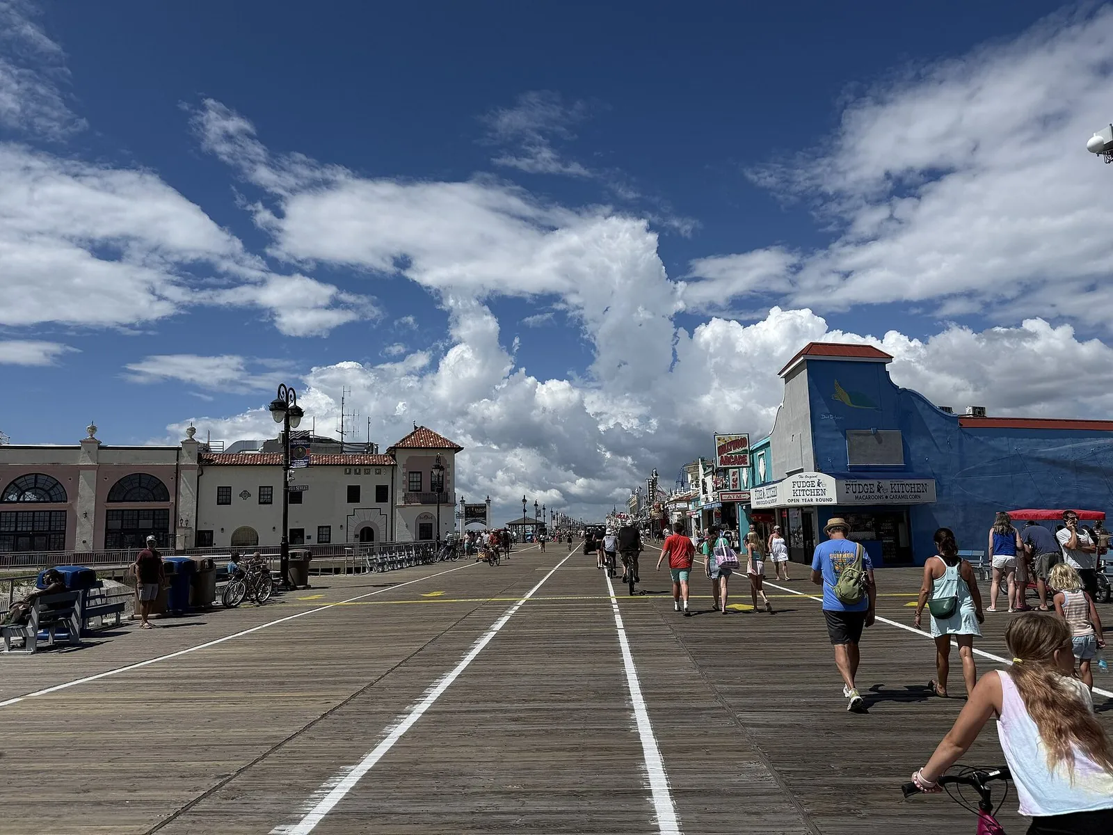The Ocean City NJ boardwalk at 8th Street on a sunny summer day, with walkers and a cyclist on the boards, a Spanish-style pavilion on the left and shops on the right
