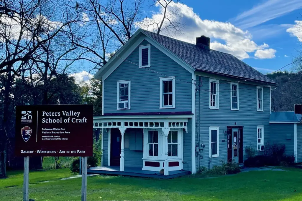 A blue clapboard farmhouse with a white porch and a Peters Valley School of Craft sign on its lawn, under a bright sky