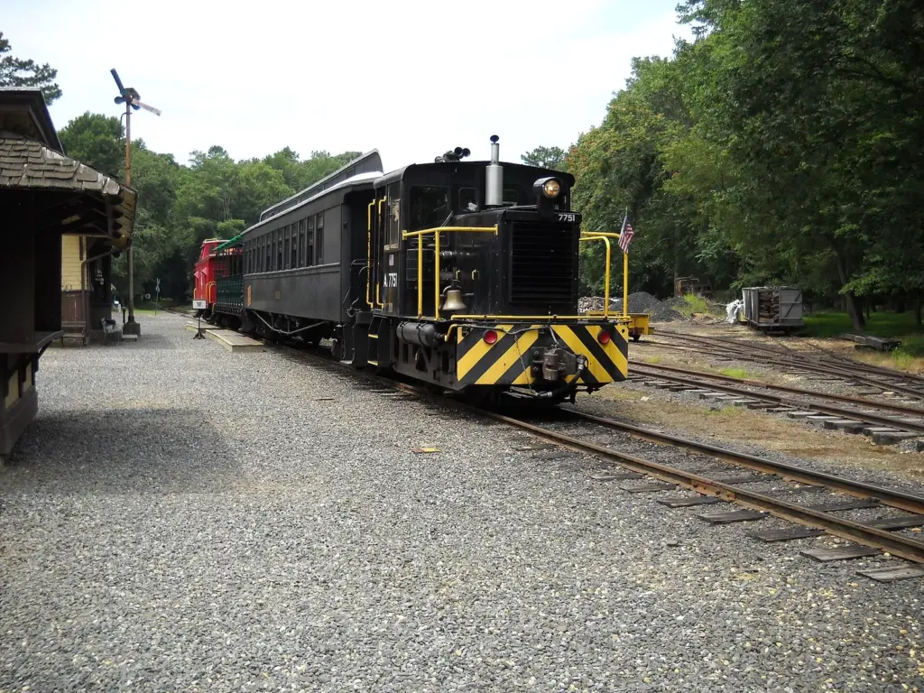 A black and yellow diesel locomotive pulling a vintage passenger car past a small wooden station on a narrow gauge railroad in the woods