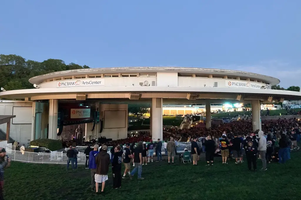 The round, white-roofed amphitheater at dusk, seen from the lawn, with a crowd standing on the grass and the stage lit under the canopy