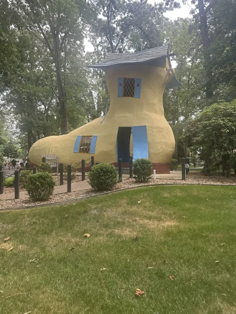 A giant yellow shoe-shaped house with blue windows and a small roof in a grassy park at Storybook Land