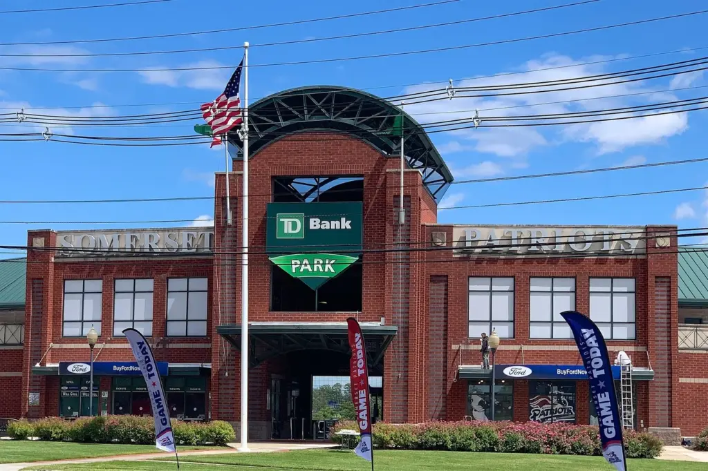 The red-brick entrance of a minor-league ballpark with a green TD Bank Park sign over the gate, an American flag and team banners out front