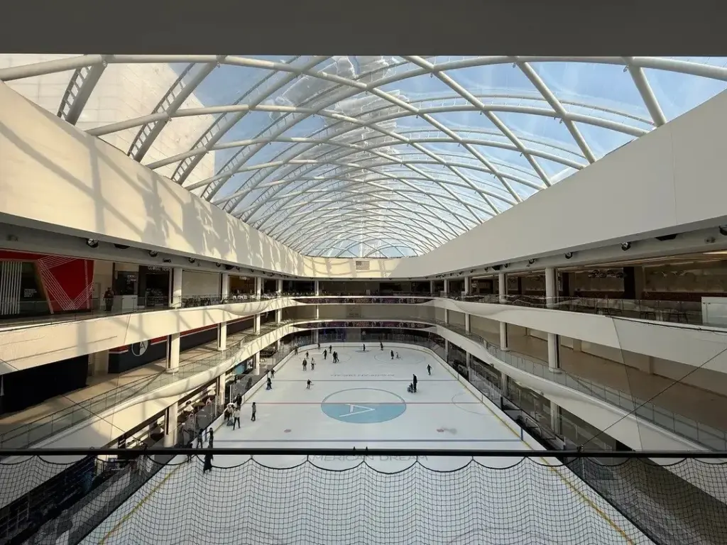 Skaters on an indoor ice rink seen from an upper level, under a long barrel-vaulted glass roof lined with shopping balconies
