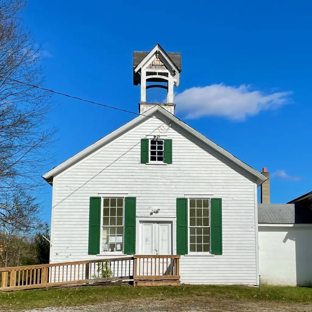 A white clapboard one-room schoolhouse with green shutters and a small bell tower at Walpack Center, under a bright blue sky